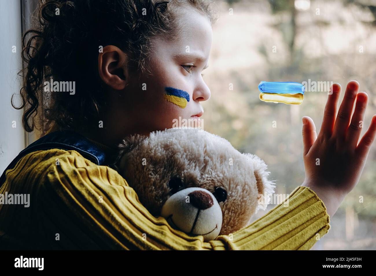 Vista laterale di triste ragazzino con bandiera di ucraina sul viso, bambino stanco che tiene morbido orso seduto da solo in windowsill a casa. Pace, nessuna guerra, hel Foto Stock