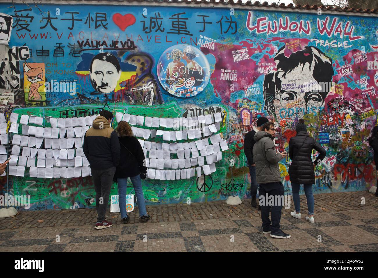 il presidente russo Vladimir Putin ha raffigurato accanto a John Lennon sul muro di Lennon (Lennonova zeď) a Praga, Repubblica Ceca. Il presidente Vladimir Putin è raffigurato con i caratteristici baffi da taglio e da spazzolino di Adolf Hitler collocati sulla sanguinosa mappa dell'Ucraina per protestare contro l'invasione russa dell'Ucraina nel 2022. L'iscrizione ceca sul capo di Putin 'Válka je vůl' (letterario significa: La guerra è un bue) è una linea di una canzone popolare della rock band ceca 'Synkopy 61'. I turisti nella foto del 2 aprile 2022 leggono i testi antguerra posti accanto al Muro di Lennon. Foto Stock