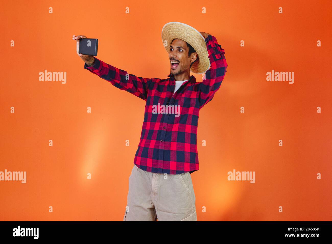 Black Man con Junina Party vestito con un tablet o cellulare isolato su sfondo arancione. Giovane uomo che indossa abiti tradizionali per Festa Junina - Foto Stock