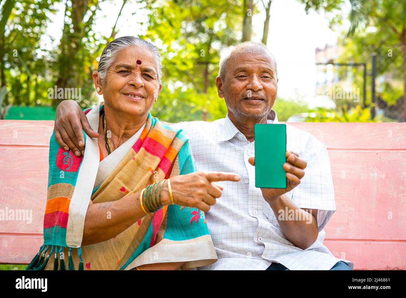 Coppia senior che mostra il telefono cellulare con schermo verde puntando il dito mentre si guarda la telecamera al parco - concetto di pubblicità e app per il settore sanitario Foto Stock