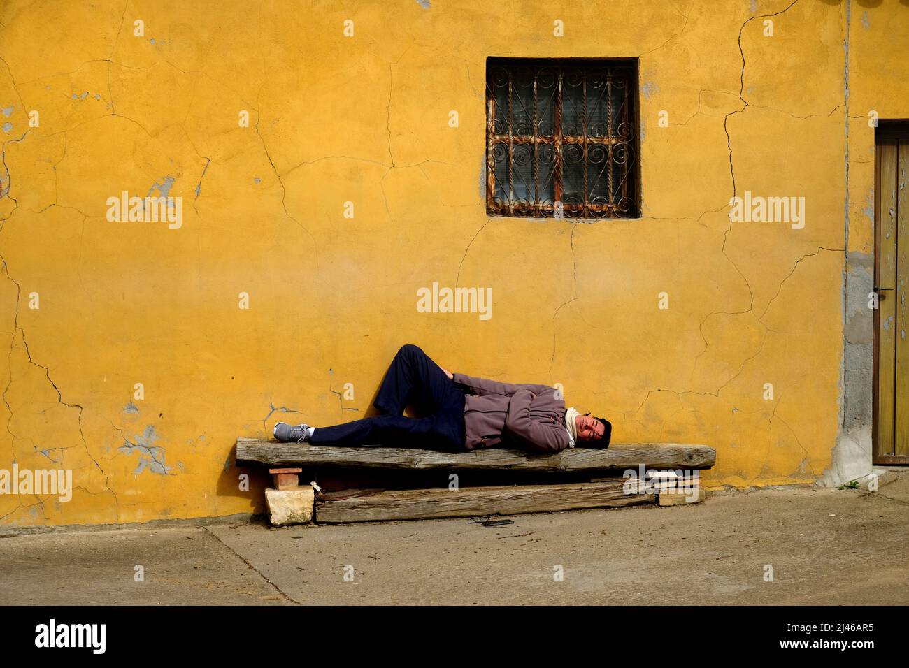 Un uomo dorme felicemente su una panca di legno ruvida al sole accanto ad un muro giallo a Becerril de Campos, vicino Palancia, Castiglia e León, Spagna Foto Stock