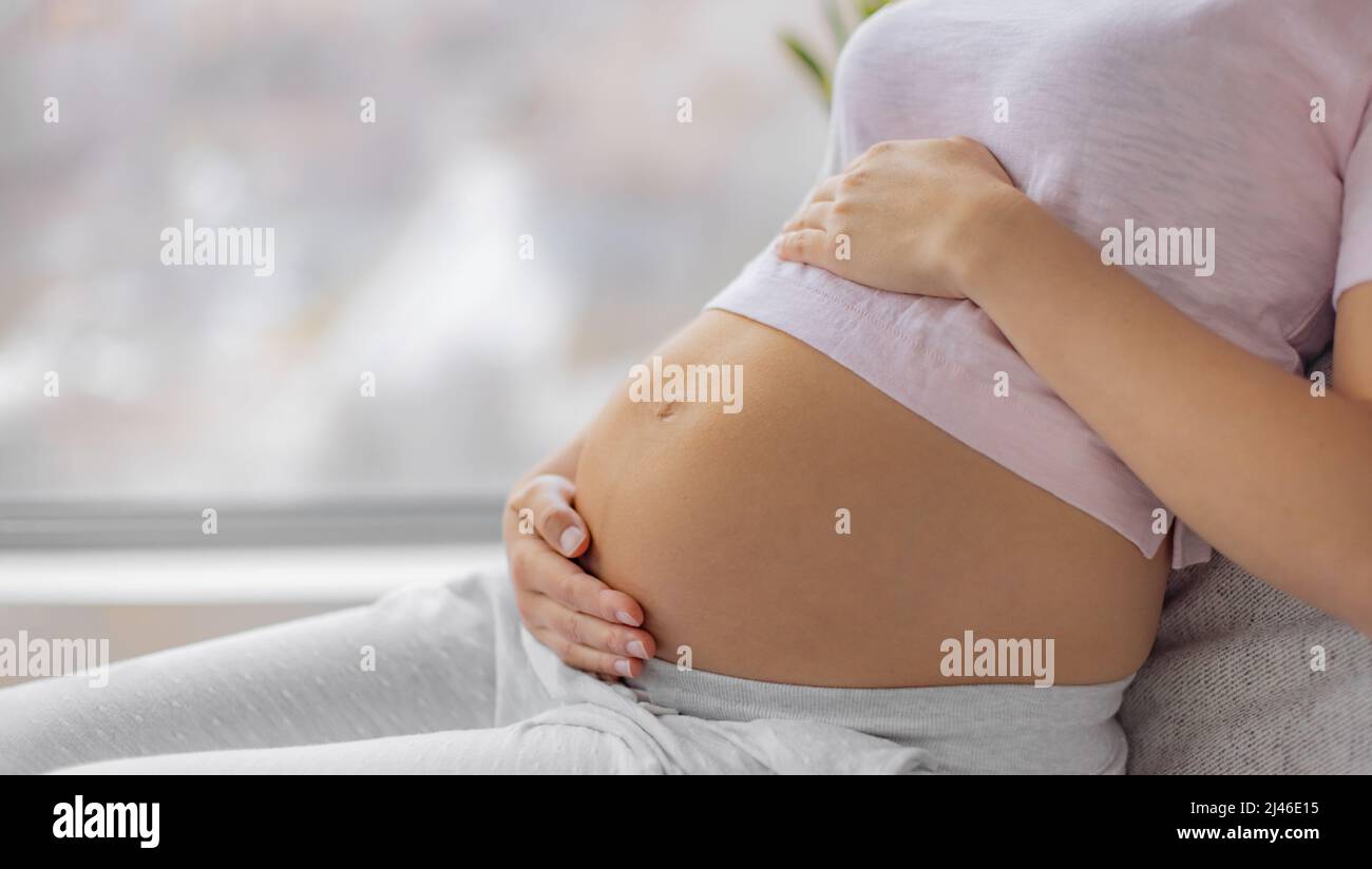 Donna incinta che si rilassa a letto in pigiama seduta sul davanzale carezzando il ventre in attesa di un bambino. Primo piano di mani che tengono lo stomaco, t-shirt rosa Foto Stock