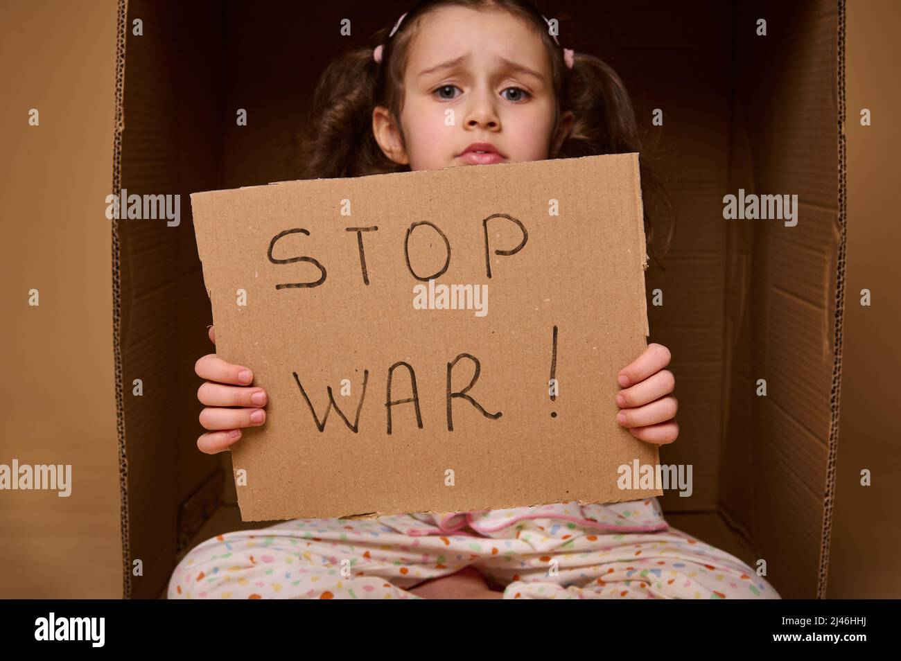 Primo piano piccola ragazza caucasica in abiti da casa seduti all'interno di una scatola di cartone con un poster STOP WAR. Il concetto di rifugiati e migranti che hanno perso la Th Foto Stock