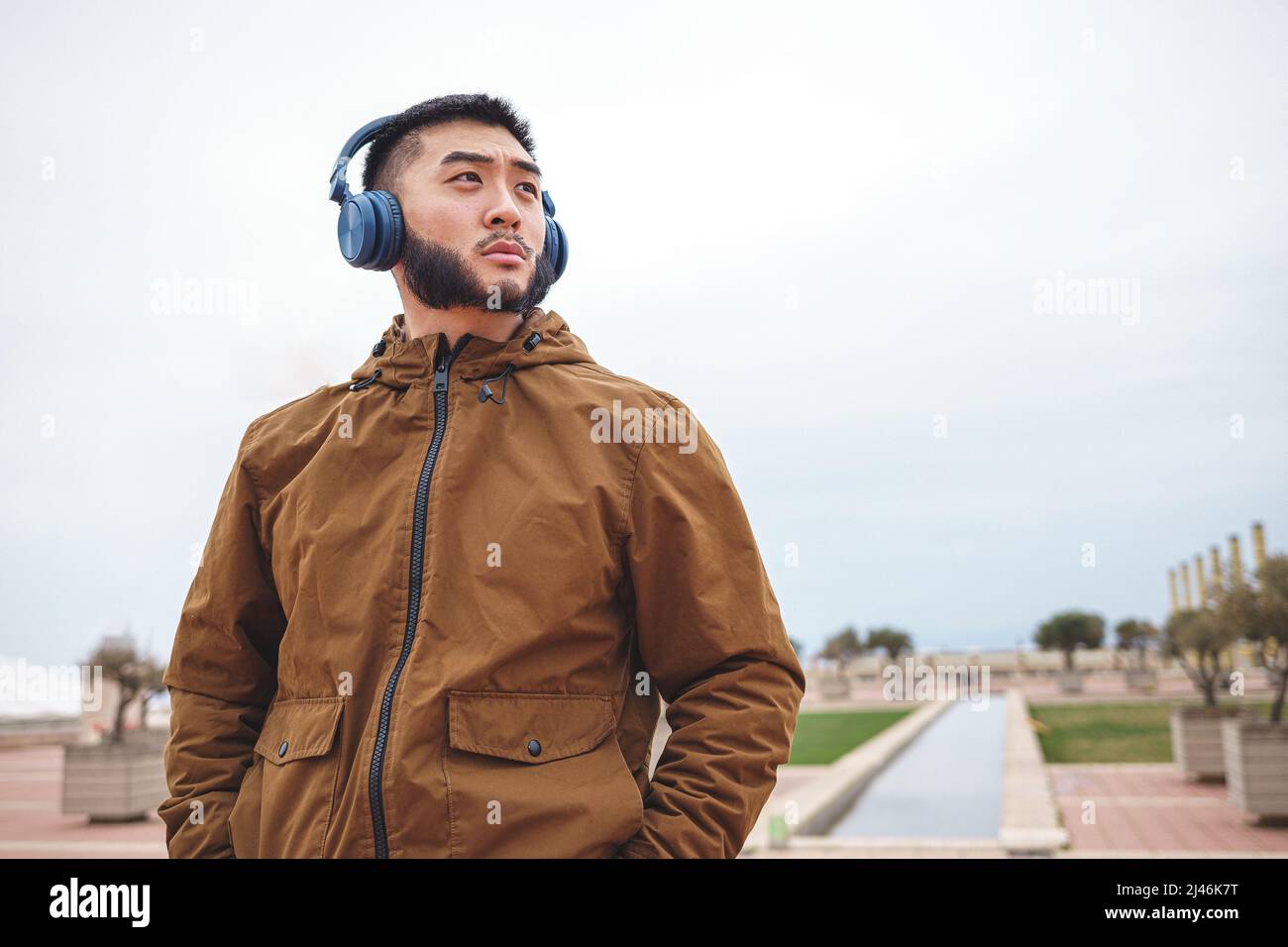 Uomo asiatico serio in outerwear ascoltare la musica nel parco Foto Stock
