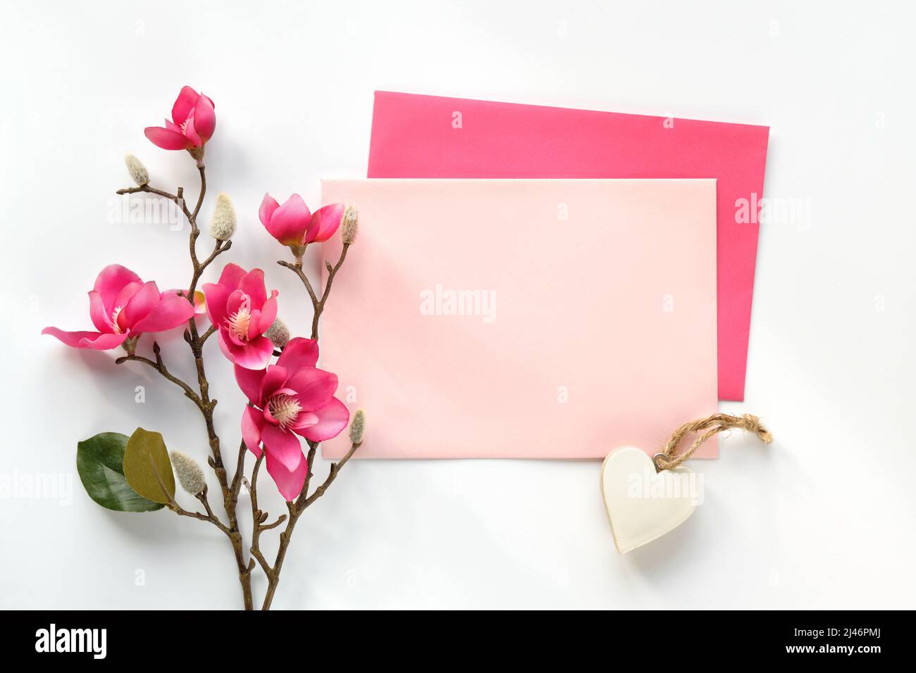Cartolina vuota con busta. Fiori decorativi rosa magnolia. Vista dall'alto su sfondo bianco. Luce solare diretta naturale, ombre. Foto Stock