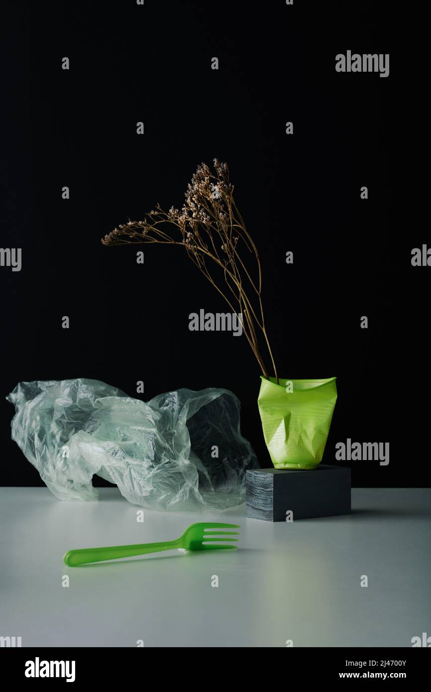Verticale concettuale STILL LIFE composizione di fiori secchi in tazza di plastica sbriciolata, sacchetto di plastica e forchetta sul tavolo, parete nera fondo Foto Stock