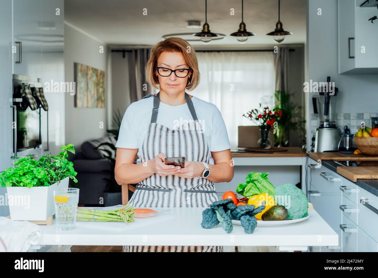 Donna che usa il telefono e cerca la ricetta online per cucinare cena in cucina a casa. Guardare il video tutorial mentre si prepara un piatto sano con il pesce Foto Stock