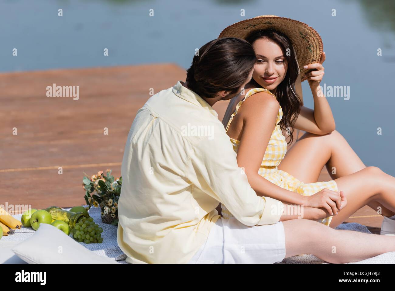 uomo che tiene le mani con donna sorridente in cappello di paglia mentre si siede sul molo durante il picnic Foto Stock