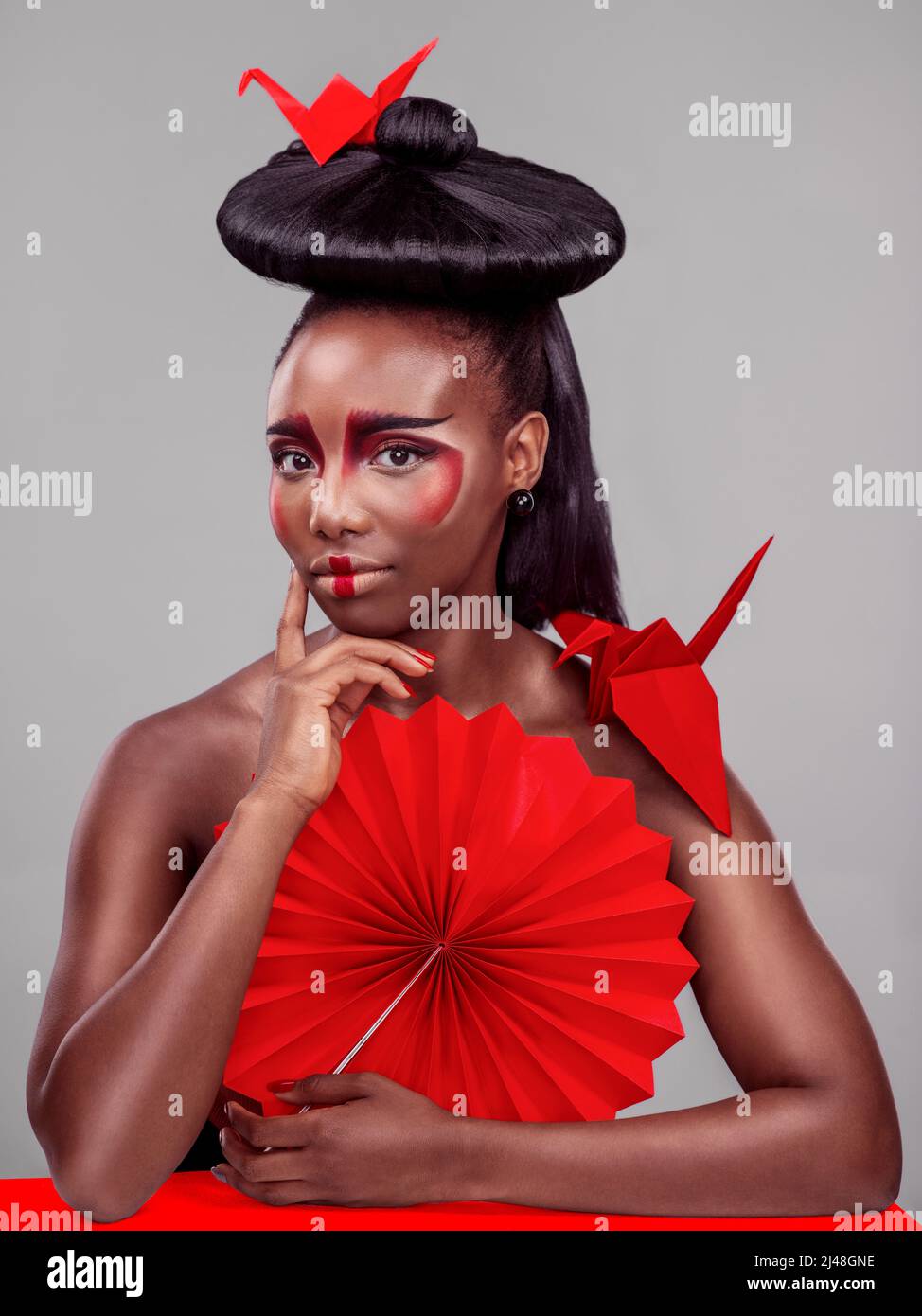 Stile africano con influenze dall'Oriente. Studio girato di una bella giovane donna che indossa trucco di ispirazione asiatica e posa con un ventilatore contro un grigio Foto Stock