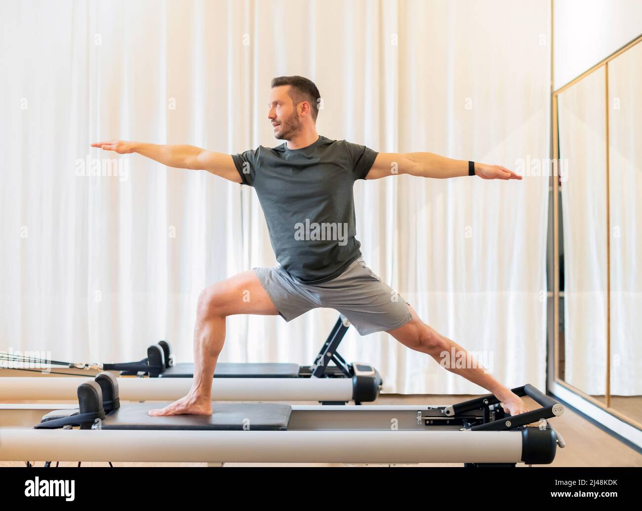 Fit uomo che esegue un affondo e stretch Warrior 2 yoga posa su un pilates riformer letto per allungare e rinforzare i suoi muscoli di anca e petto e promuovere s Foto Stock