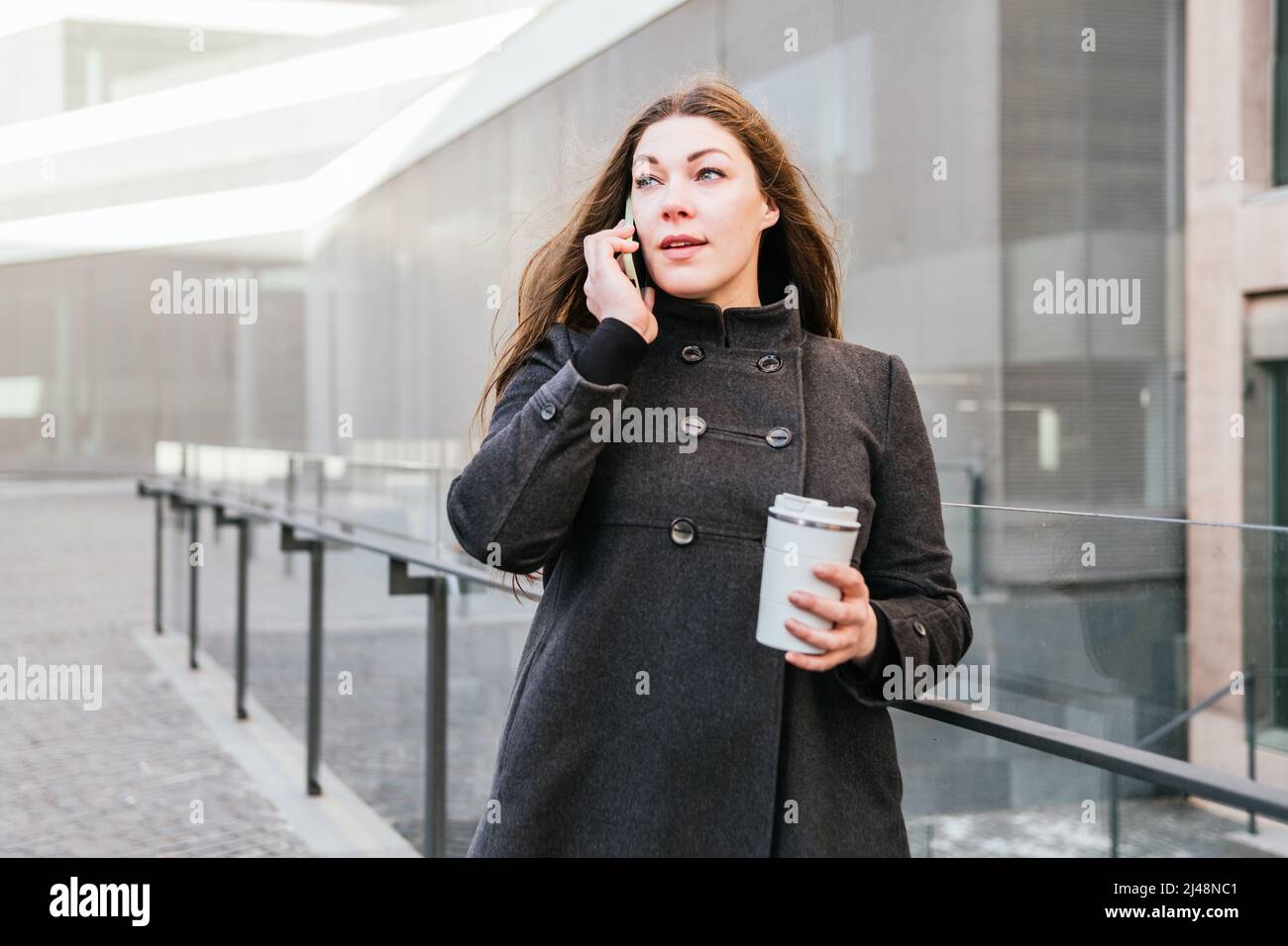 Donna indossando caldo cappotto in piedi con riutilizzabile in acciaio inox Tumbler per bevanda calda vicino vetro recinto e parlare sul cellulare vicino ufficio edificio Foto Stock