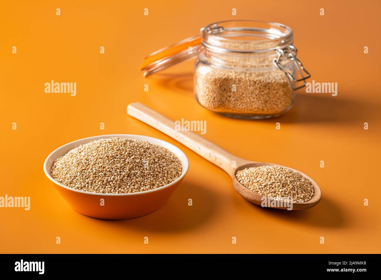 Semi bianchi di quinoa in una ciotola, cucchiaio di legno e clip top vaso di vetro su sfondo arancione brillante. Cottura chenopodium quinoa concetto. Chicchi di quinoa crudi Foto Stock