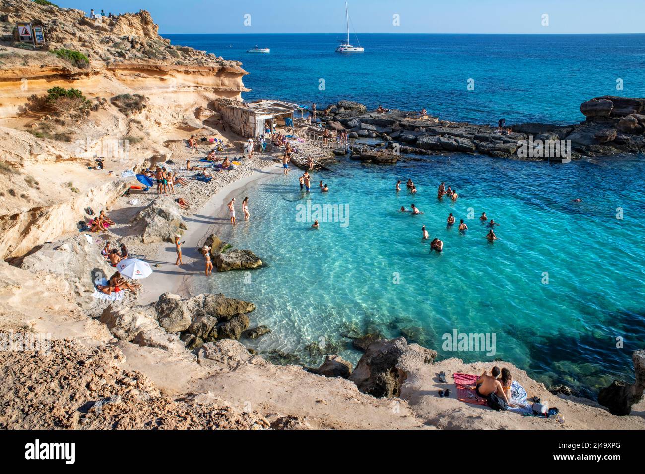 Es Caló d´es Mort cala spiaggia, i visitatori estivi adagiati su un'insenatura fiancheggiata da scogliere rosse e rocce in acque turchesi, Formentera Balearic isola Foto Stock