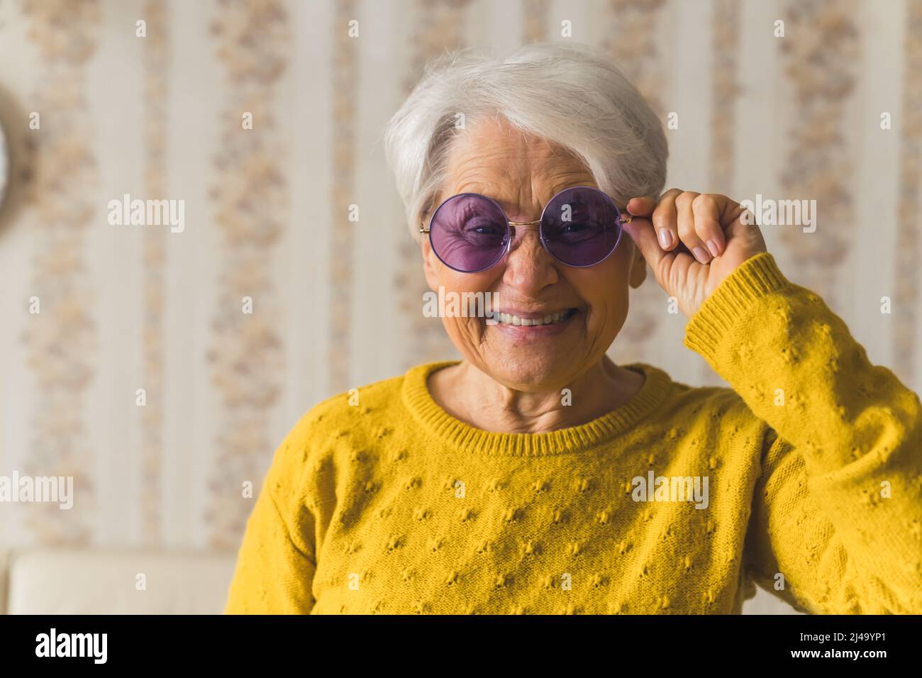 Funky divertente caucasica grigio-capelli ritirati nonna guardando e sorridendo alla macchina fotografica mentre indossano moderno occhiali violetto. Scatto medio al coperto con carta da parati vintage. Foto di alta qualità Foto Stock