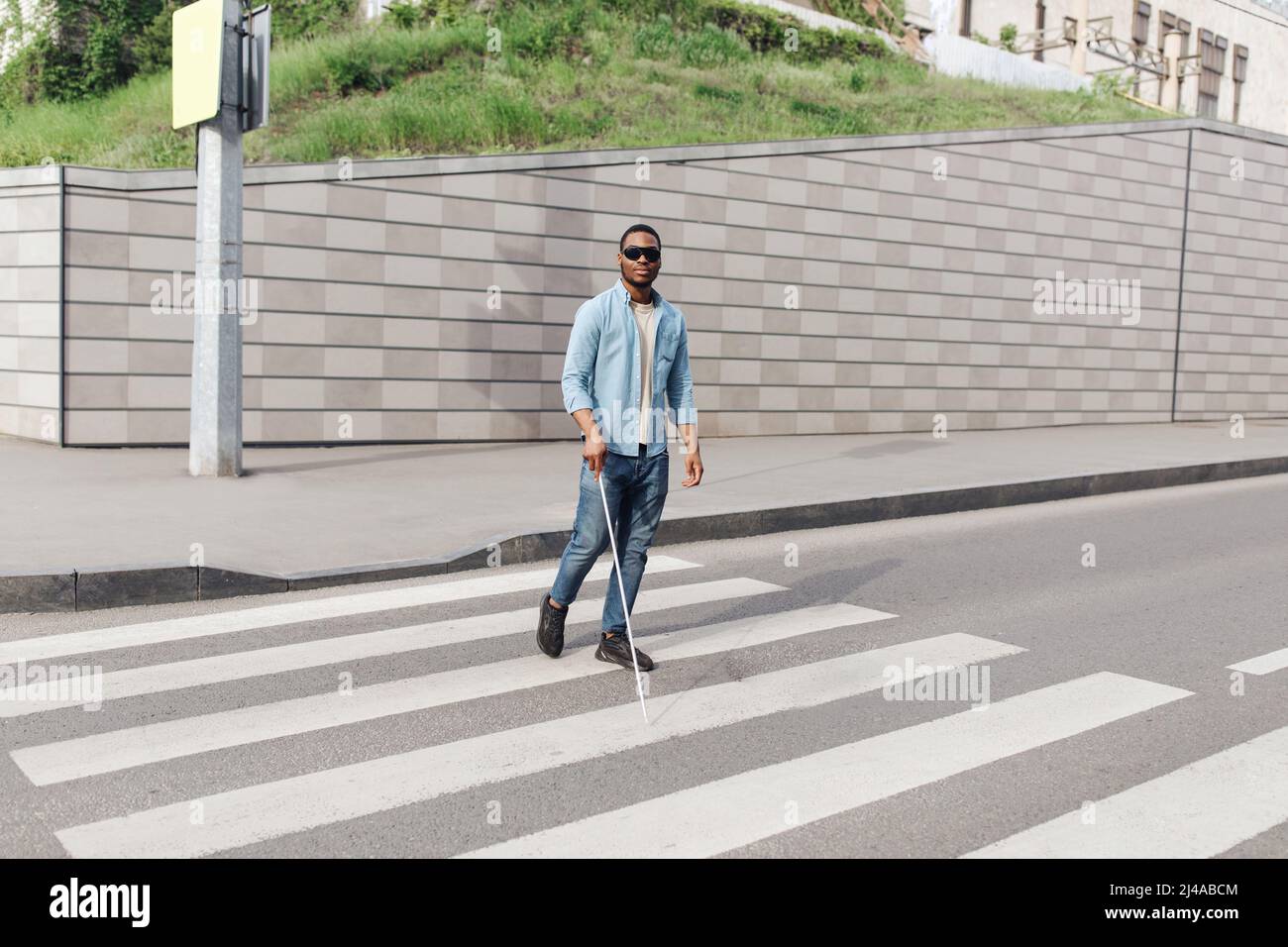 Giovane uomo nero con disabilità visiva indossando occhiali scuri, usando bastone da passeggio per attraversare la strada nel centro della città Foto Stock