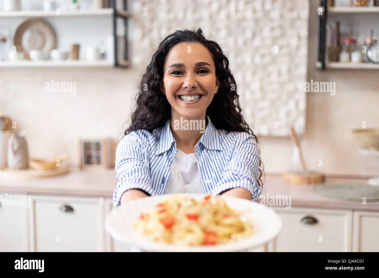 Felice donna latina che mostra spaghetti nel piatto, allungando il pasto alla macchina fotografica e sorridendo Foto Stock