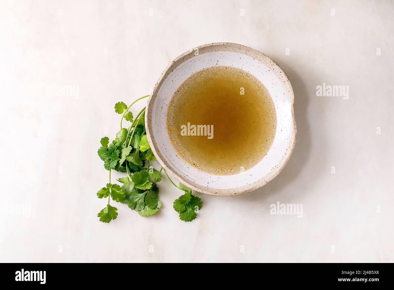 Brodo di pollo o anatra chiaro fatto in casa in ciotola di ceramica con mazzo di coriandolo verde fresco su tavolo di marmo bianco. Disposizione piatta, spazio di copia Foto Stock