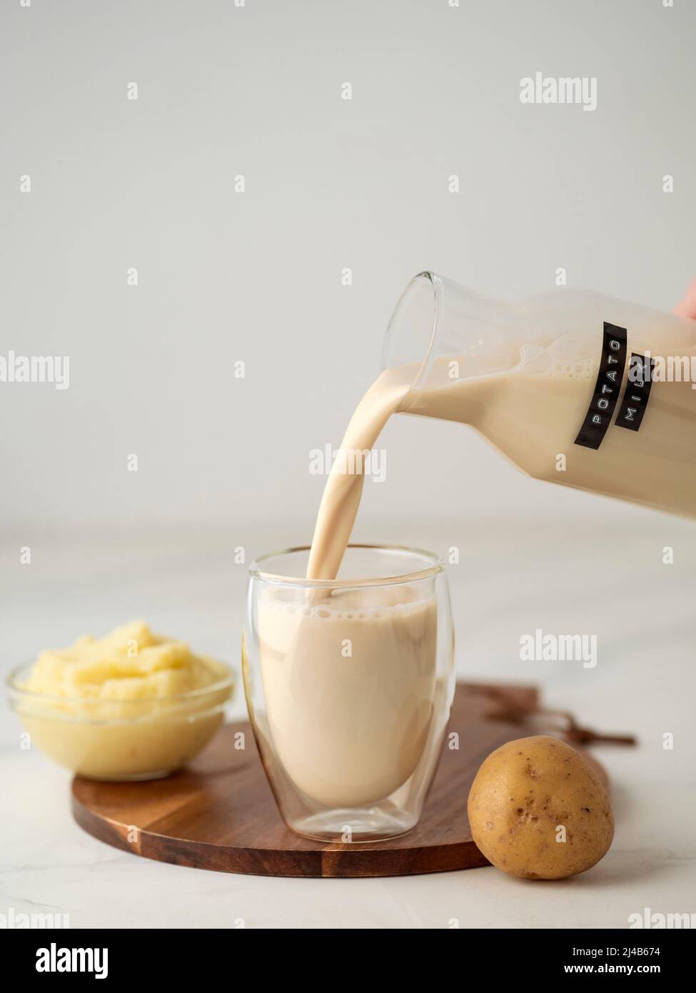 Latte di patate versato in vetro su sfondo di marmo bianco. Versare il latte vegano in vetro, con purea di patate e tuberi di patate sullo sfondo. Spazio di copia. Foto Stock