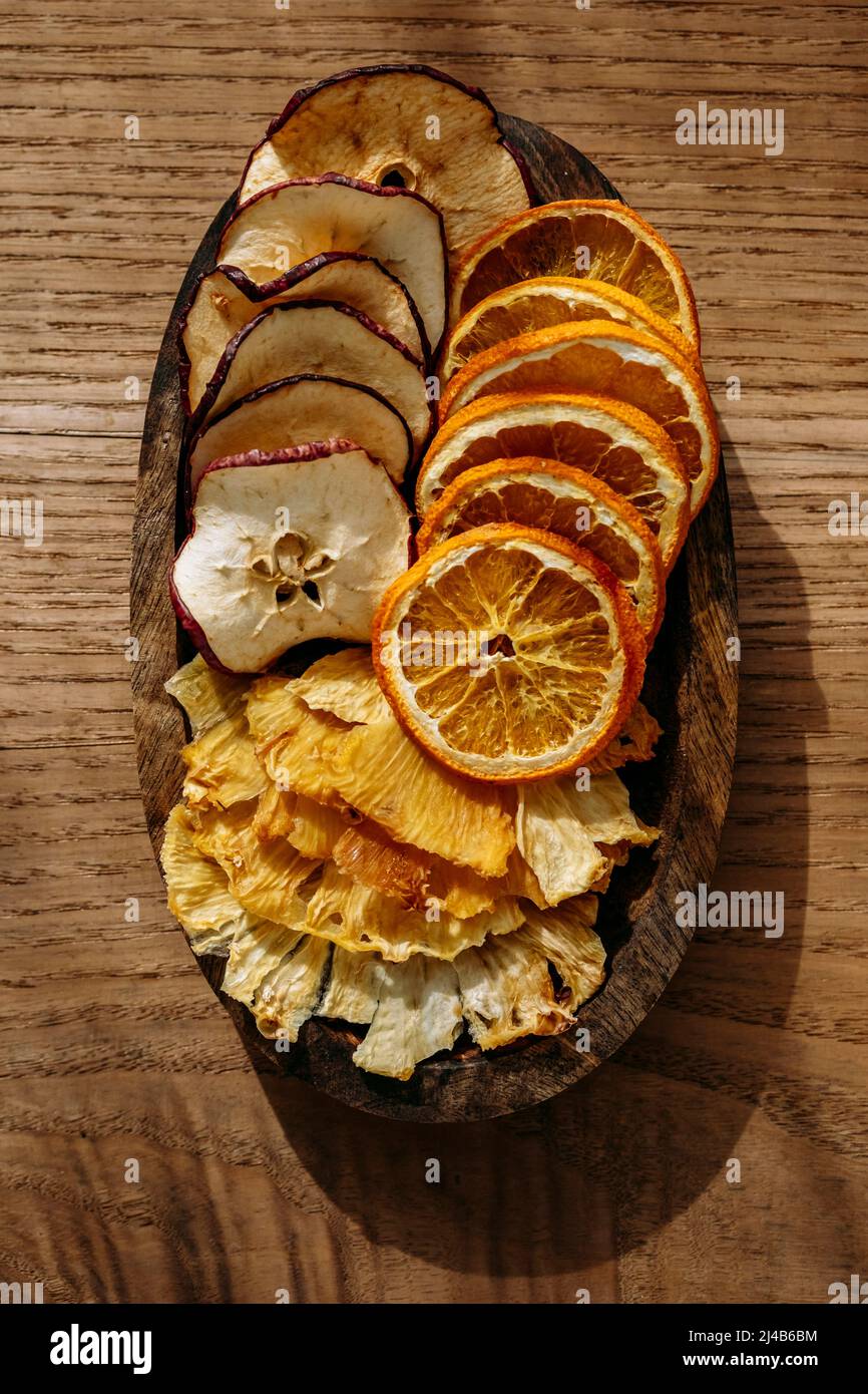 Vista dall'alto di sottili fette di frutta secca, ananas, mela e arancia, su un piccolo vassoio di legno, su un tavolo di legno Foto Stock