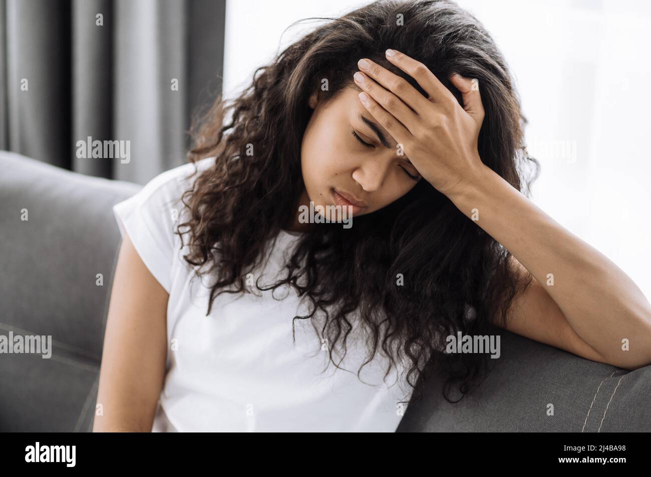 La giovane donna stanca è seduta sul divano, avendo un mal di testa. Infelice femmina afroamericana è malsana, stressata, turbata per la malattia o ha problemi Foto Stock