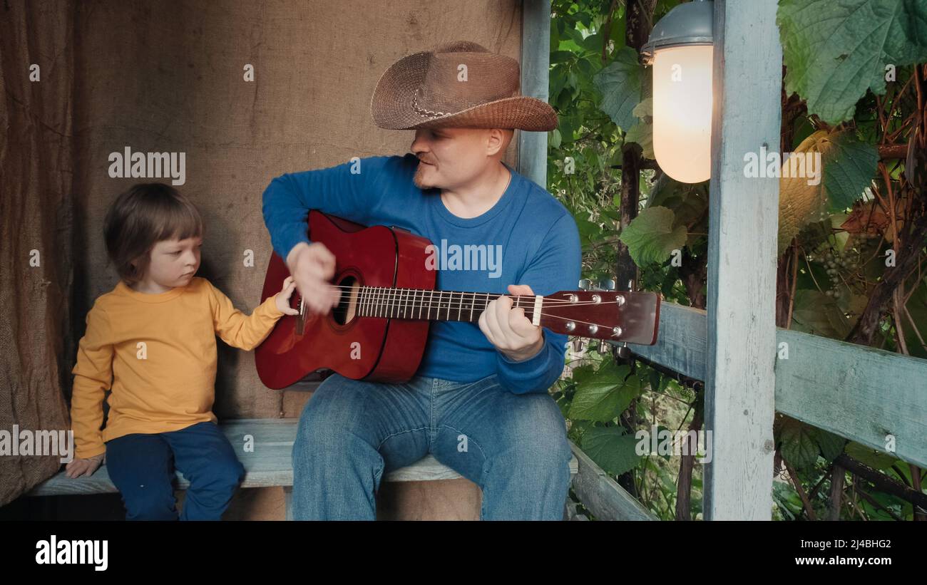 Papà cowboy suona una chitarra acustica per il suo bambino piccolo. Vecchio, arredamento retrò Foto Stock