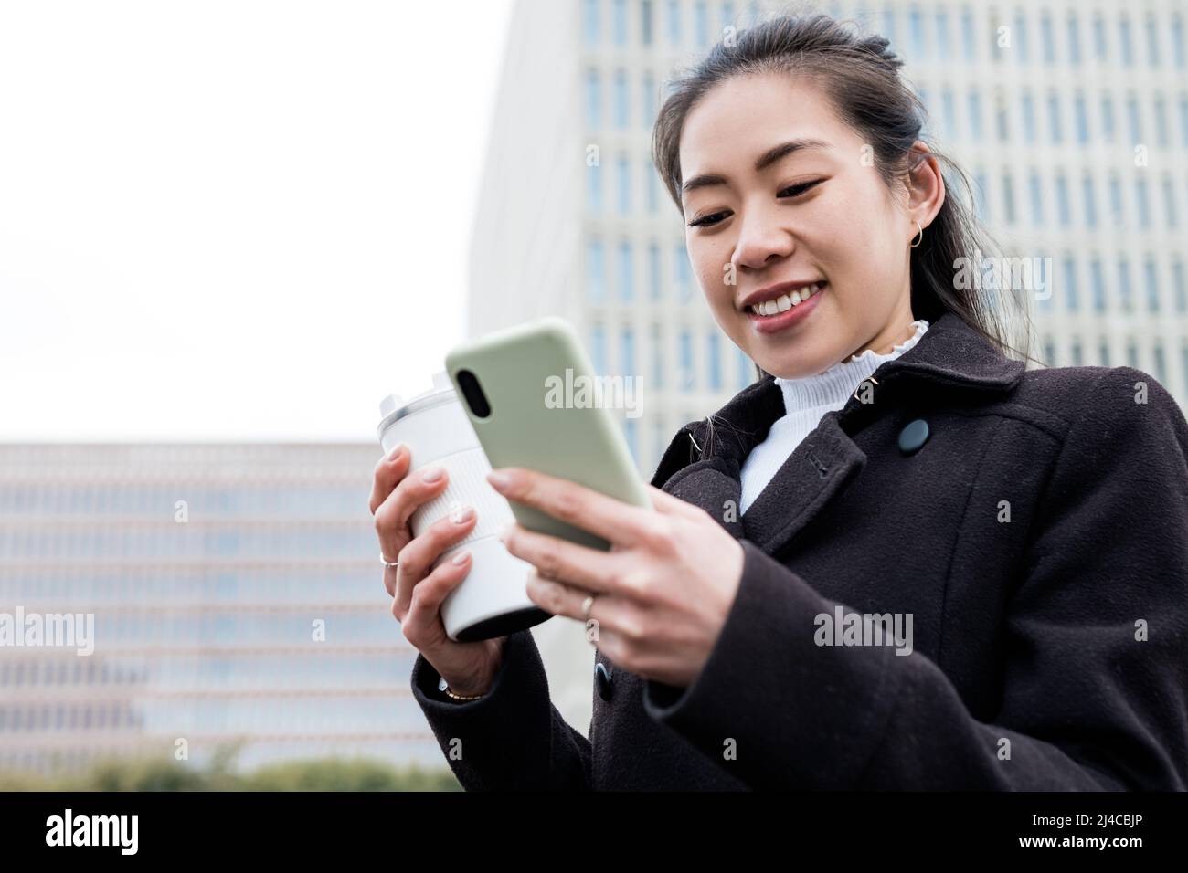 Da sotto felice imprenditore asiatico femminile in cappotto nero con eco amichevole tazza di caffè per andare. Sorride e naviga sui social media sul cellulare mentre si trova in città al mattino. Foto Stock