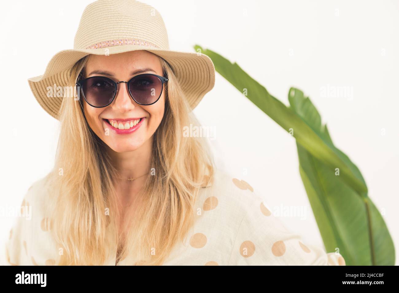 Concetto di viaggio vacanze estive. Donna bionda caucasica sorridente in un cappello e occhiali da sole grandi sorride alla macchina fotografica mentre si alza di fronte a una foglia verde sfocata su sfondo bianco. Foto di alta qualità Foto Stock