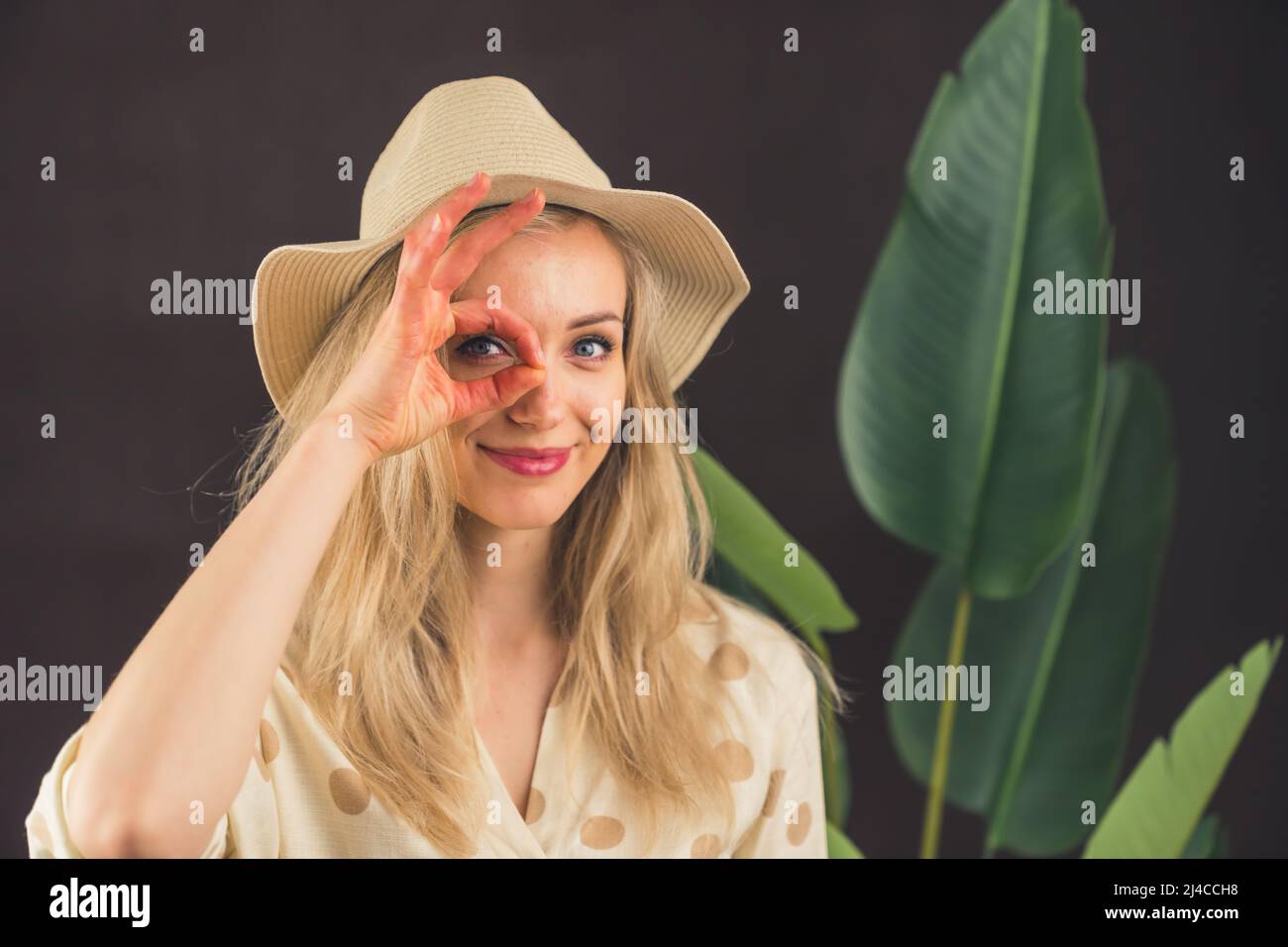 Divertente ragazza bionda caucasica in piedi di fronte a una pianta a foglia grande in un cappello che fa un gesto ok con mano sorridente, occhio guardando attraverso le dita con volto felice. Concetto di viaggiatore. Safari. Foto di alta qualità Foto Stock