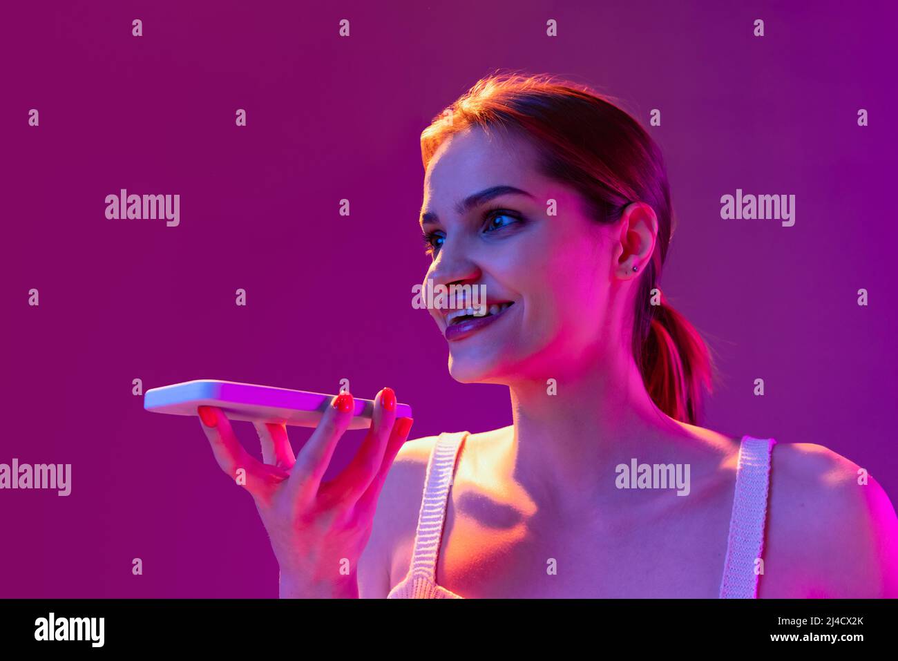 Closeup giovane bella ragazza felice registrazione messaggio vocale sul telefono isolato su sfondo viola in filtro neon. Concetto di emozioni, digitale Foto Stock
