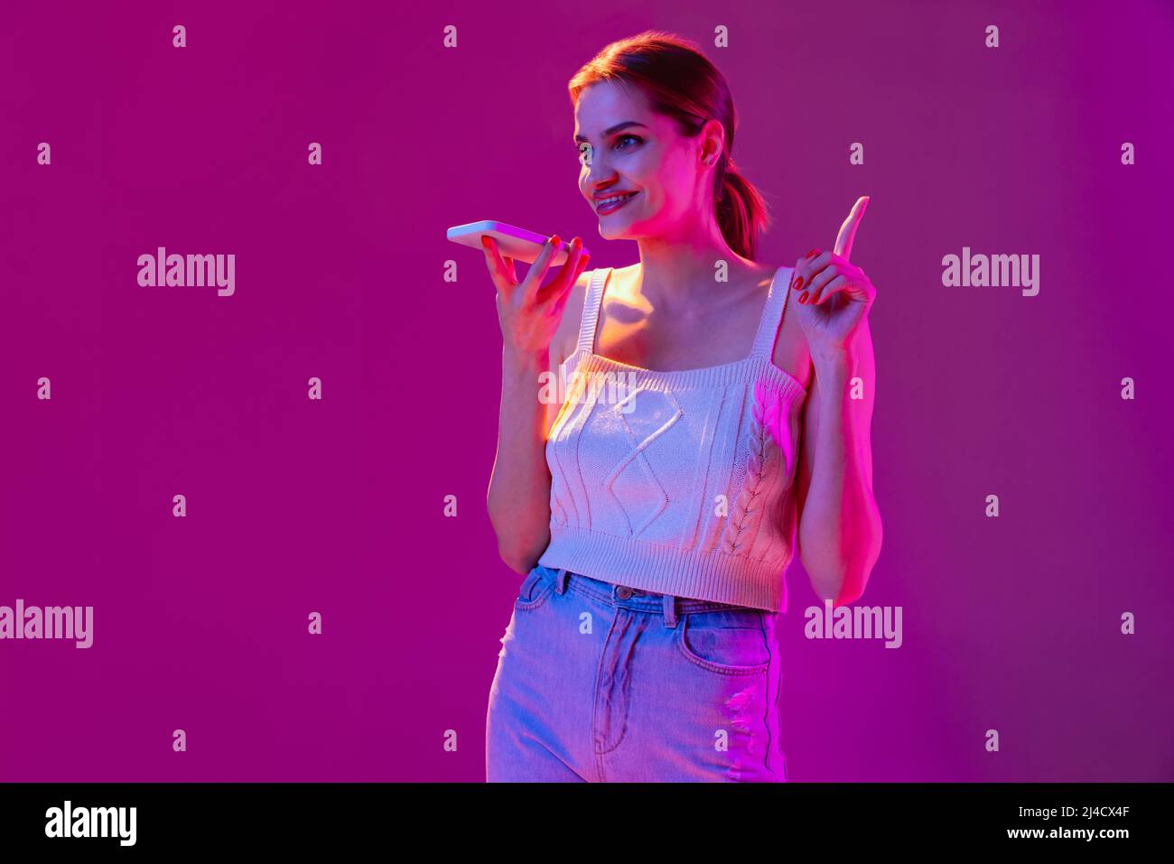 Giovane bella ragazza felice registrazione messaggio vocale sul telefono isolato su sfondo viola in filtro neon. Concetto di emozioni, tecnologie digitali Foto Stock