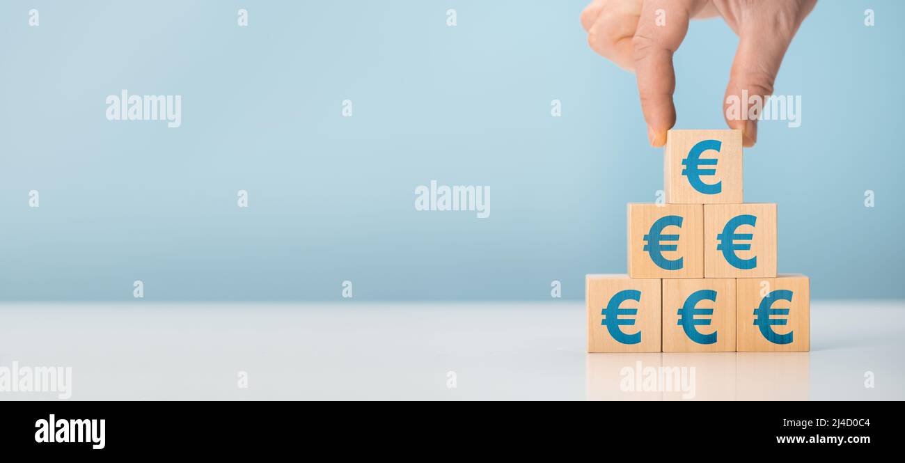 crescita delle entrate. l’uomo d’affari raccoglie la piramide dell’euro su cubi di legno su sfondo blu. Icone finanziarie o di sviluppo di affari. Dollaro su legno Foto Stock