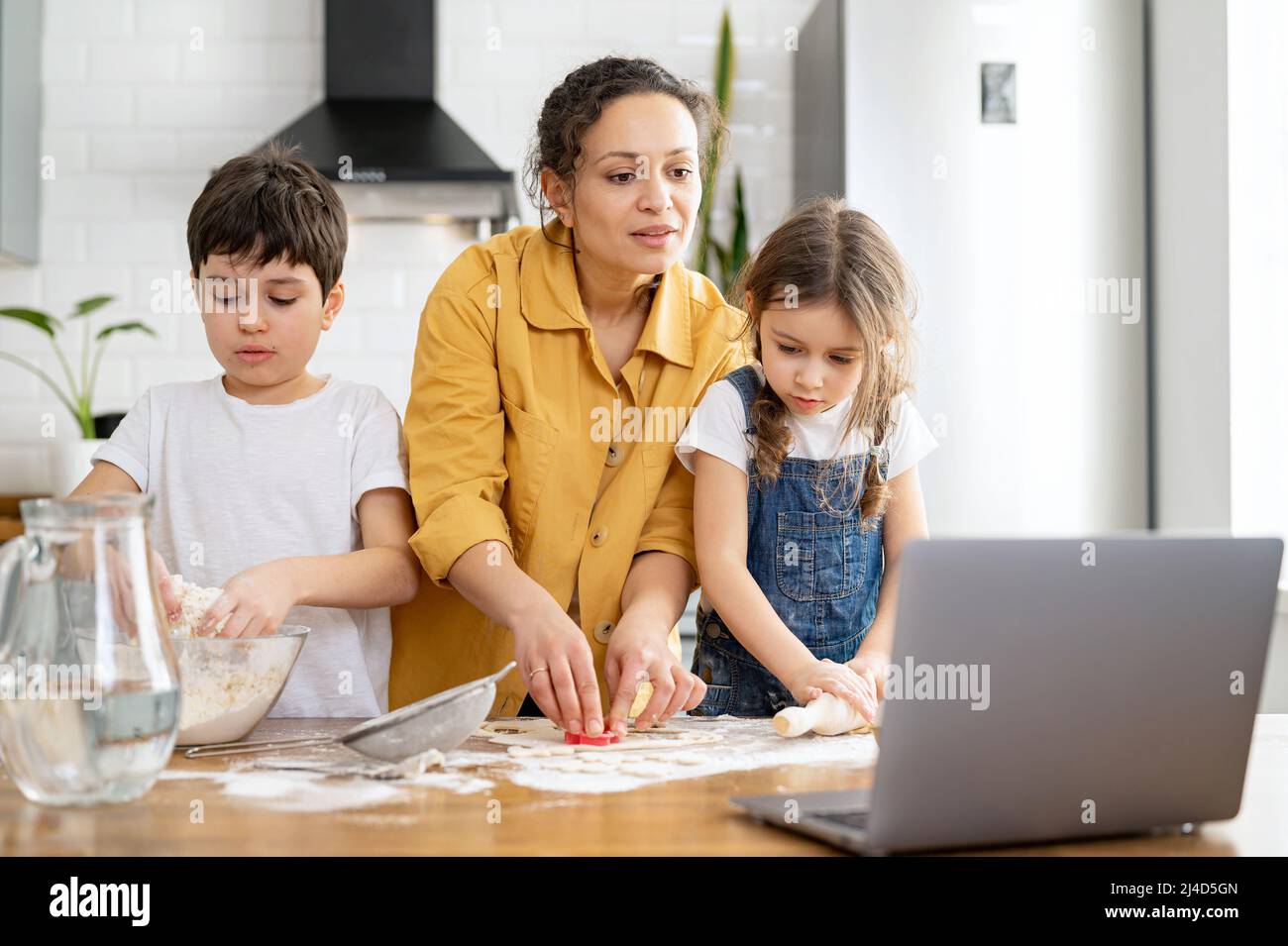 La madre di mezza età e i suoi figli che cucinano insieme, impastano l'impasto, utilizzando il matterello. Mamma e bambini trascorrono il tempo libero a casa, cuocere i biscotti, guardare un tutorial sul laptop, godersi le attività del fine settimana Foto Stock