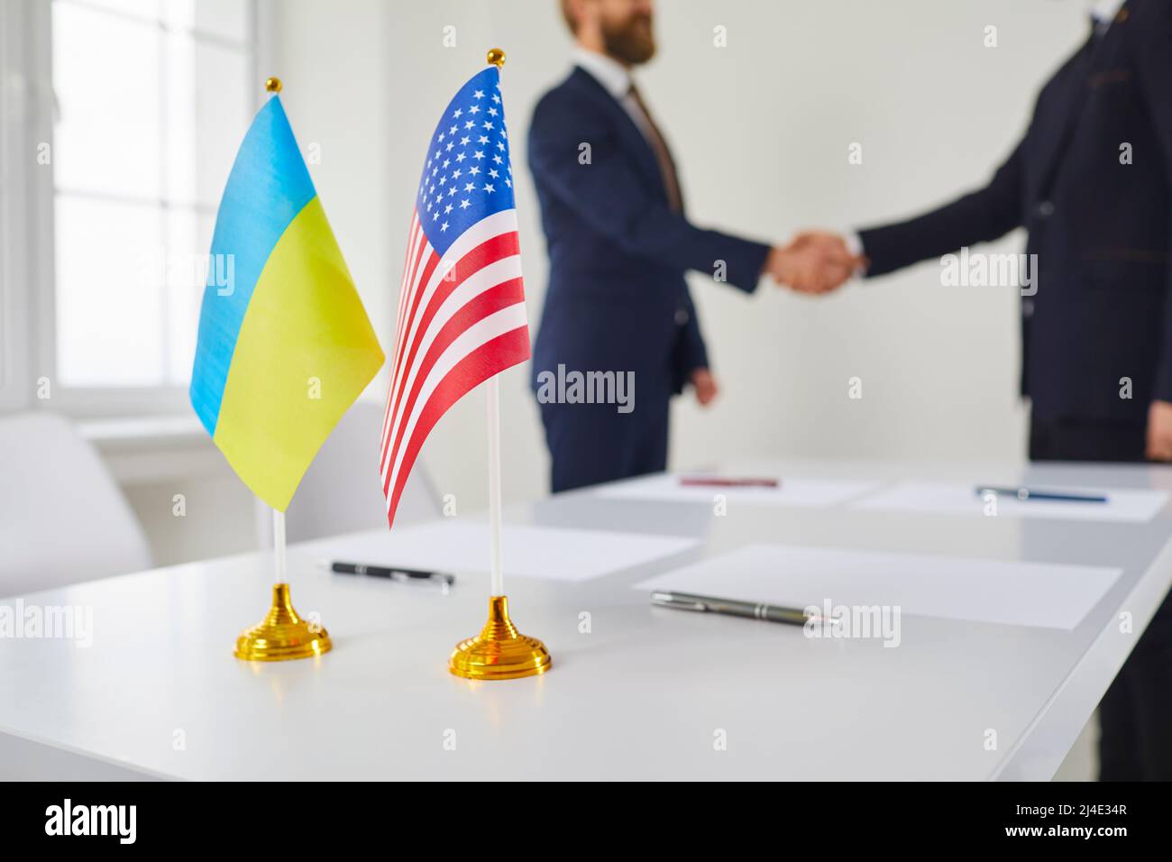 Bandiere di Ucraina e Stati Uniti sul tavolo con due uomini in background scambiano handshake Foto Stock