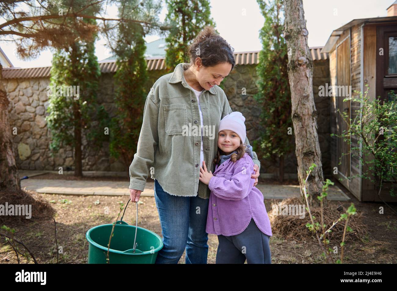Felice ragazza caucasica che abbraccia la sua madre amorevole mentre innaffia le piante nel cortile della loro casa di campagna, in piedi insieme nel giardino e. Foto Stock