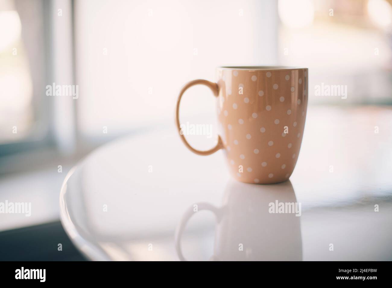 Tazza di caffè rosa con pois polka su un tavolo bianco che riflette la luce diurna della finestra. Una tazza solitaria Foto Stock Tazza di caffè rosa con pois polka su un tavolo bianco che riflette la luce diurna della finestra. Una tazza solitaria Foto Stock