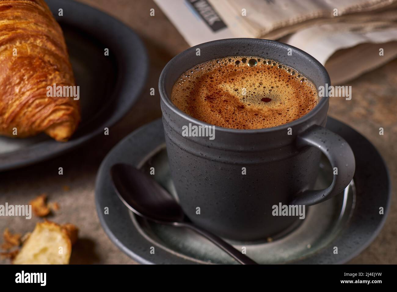Caffè nero con schiuma in tazza scura e croissant sul tavolo Foto Stock