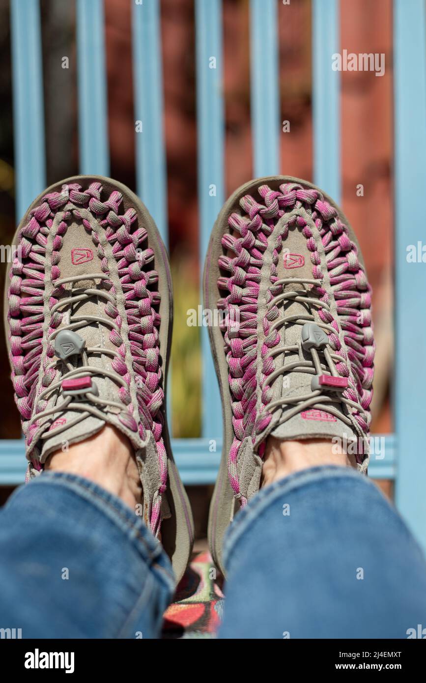 Primo piano di vitelli da donna in jeans e piedi con scarpe Uneek aceite poggiate sul sedile di sedia, le doghe di legno blu dello schienale della sedia riempiono lo sfondo. Foto Stock