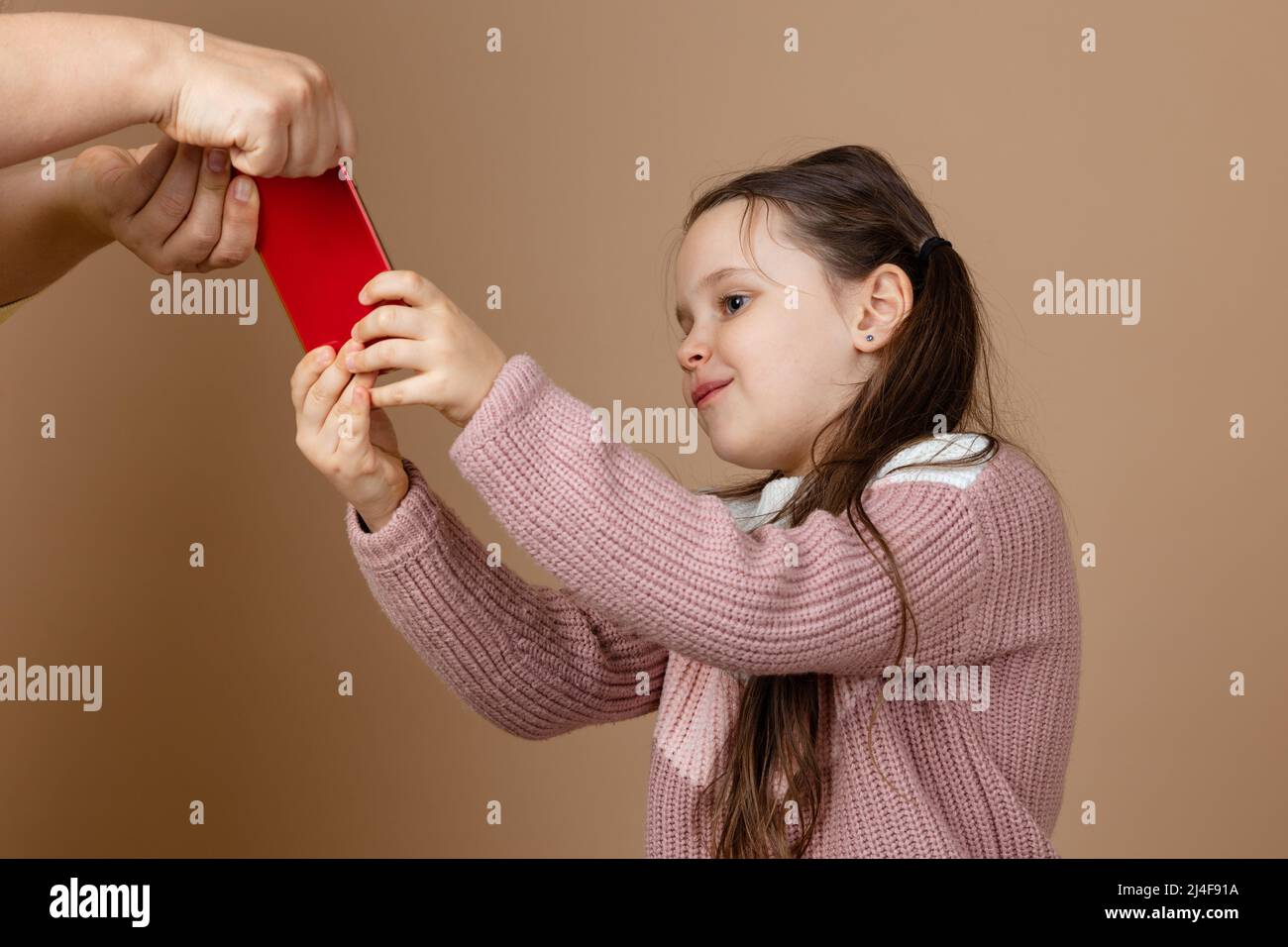 Ritratto del genitore che porta lo smartphone fuori dalle mani delle ragazze, sfondo beige. Figlia felice tenere il telefono e combattere indietro. Concetto di prevenzione di Foto Stock