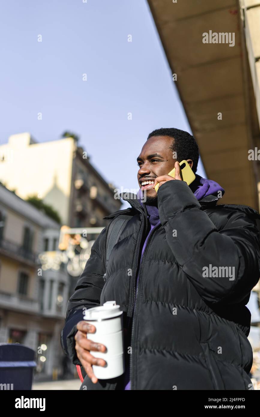 L'uomo afro-americano a basso angolo è in outerwear casual con una tazza di caffè ecologica da portare sempre con te. Guarda via con un sorriso e risponde alle chiamate telefoniche il giorno di sole nella città moderna Foto Stock