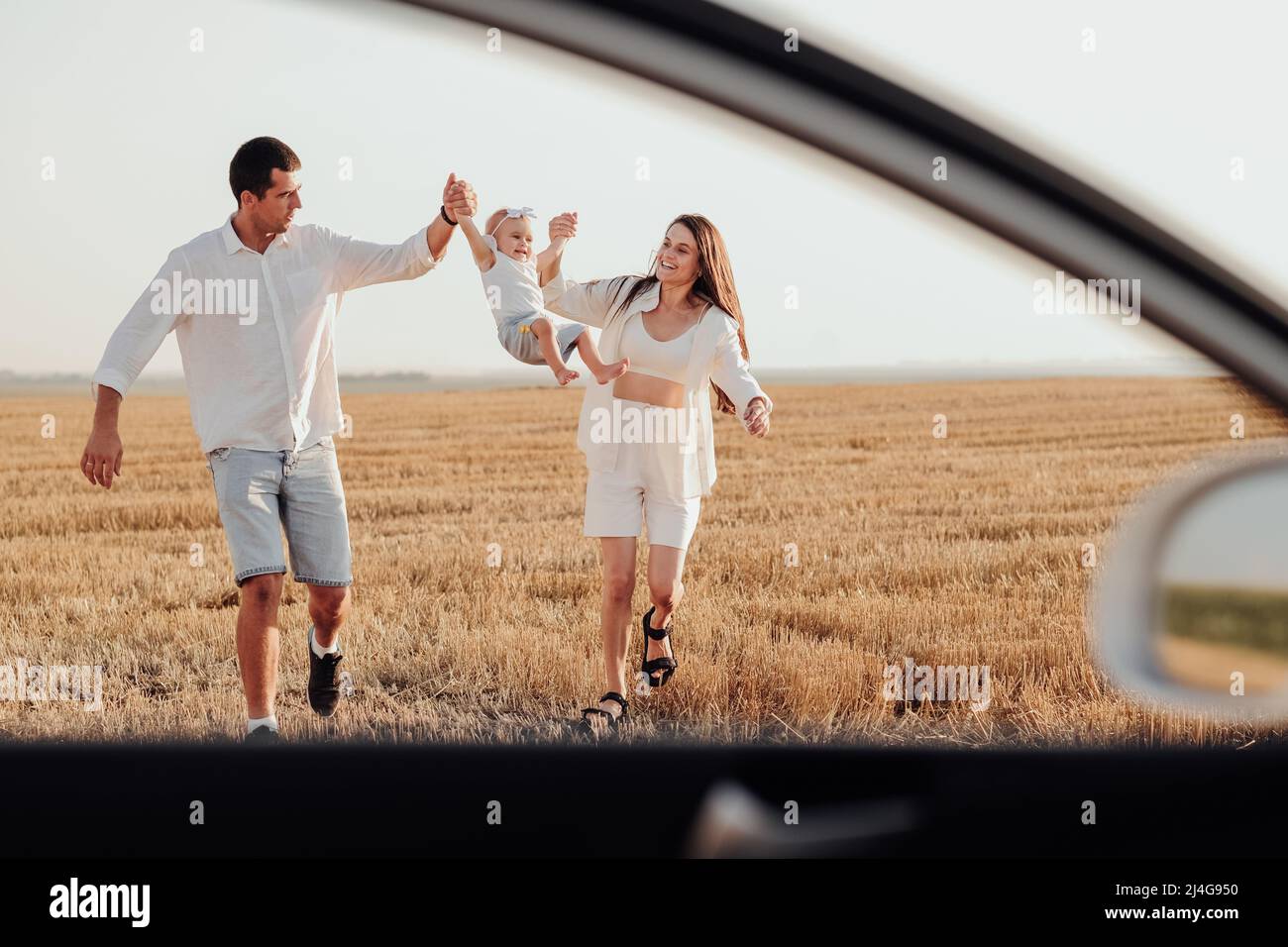 Ammira la finestra dell'auto, la mamma e il papà che tiene la loro figlia al tramonto mentre cammina sul campo Foto Stock