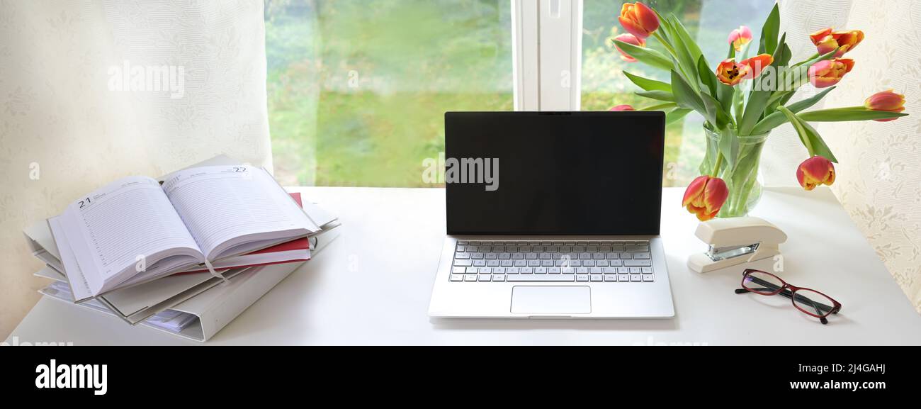 Computer portatile con schermo nero vuoto su una scrivania bianca di fronte alla finestra, anche cartella, organizer e tulipano bouquet, mock up per studio o casa d'affari, p Foto Stock