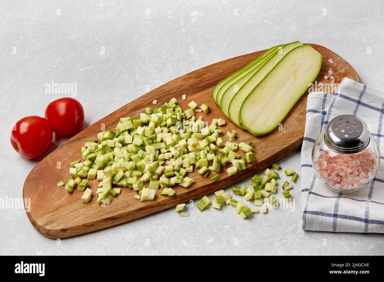 Affettato zucchine verdure zucchine su un tagliere di legno, pomodori, sale rosa e tovagliolo su un tavolo chiaro di cemento. Ingredienti per insalata di verdure Foto Stock