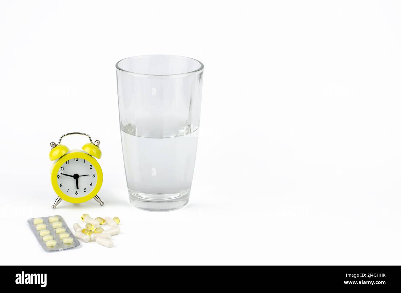 Un bicchiere d'acqua, una sveglia gialla e pillole su sfondo bianco Foto Stock