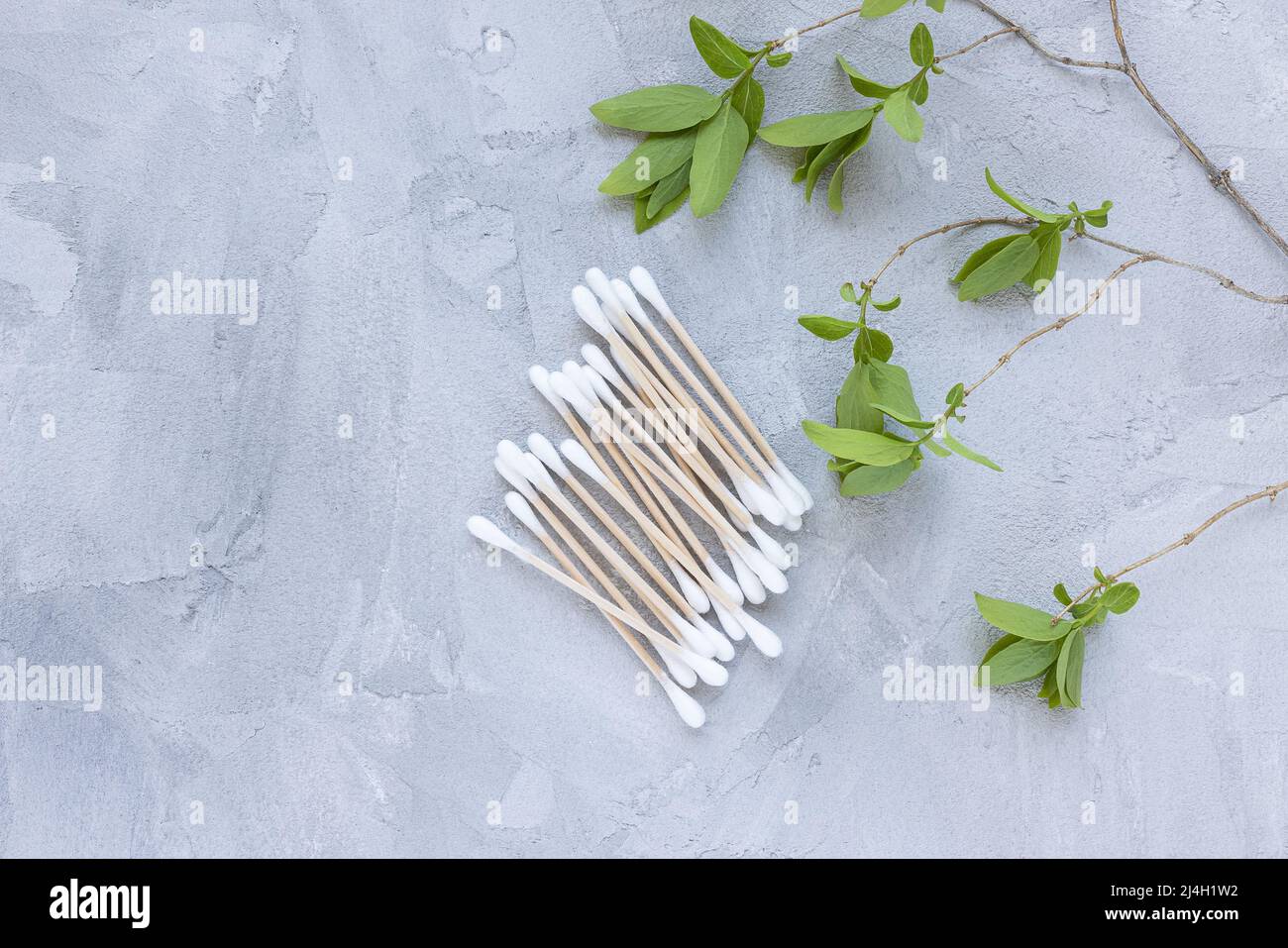 Germogli in cotone di bambù ecologico su sfondo grigio con foglie verdi. Tamponi in cotone alla moda, bastoncini di bambù per la pulizia delle orecchie. Disposizione piatta, vista dall'alto, spazio di copia Foto Stock