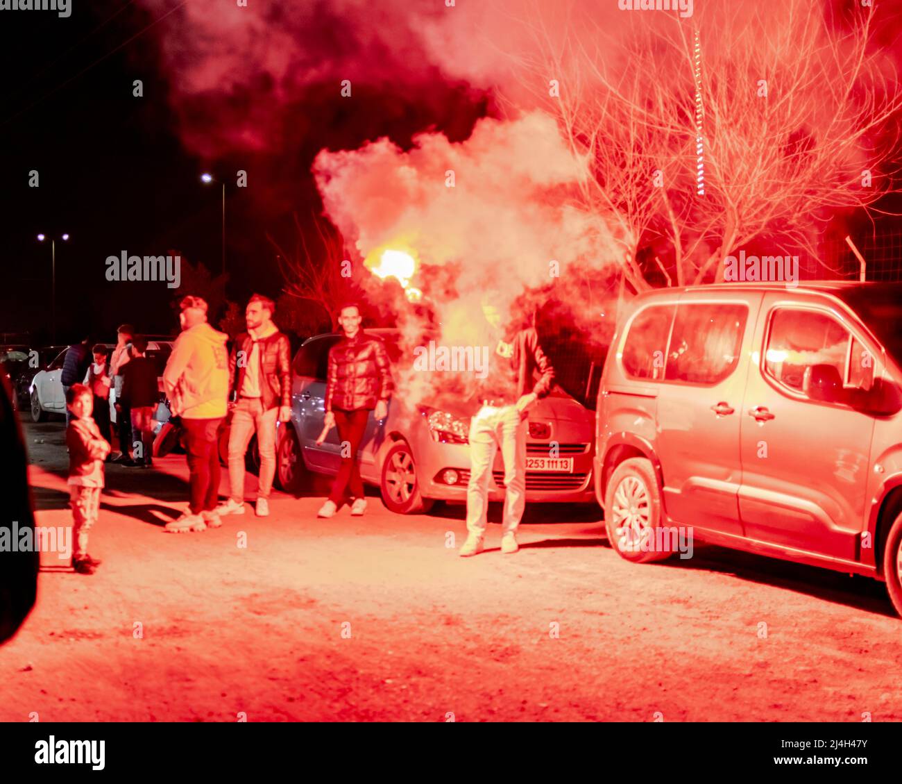 Un gruppo di giovani ragazzi che giocano con una granata di fumo rosso di notte, fumoso vapore di haze. Auto parcheggiate e un ragazzo che tiene la granata con il braccio in aria. Foto Stock
