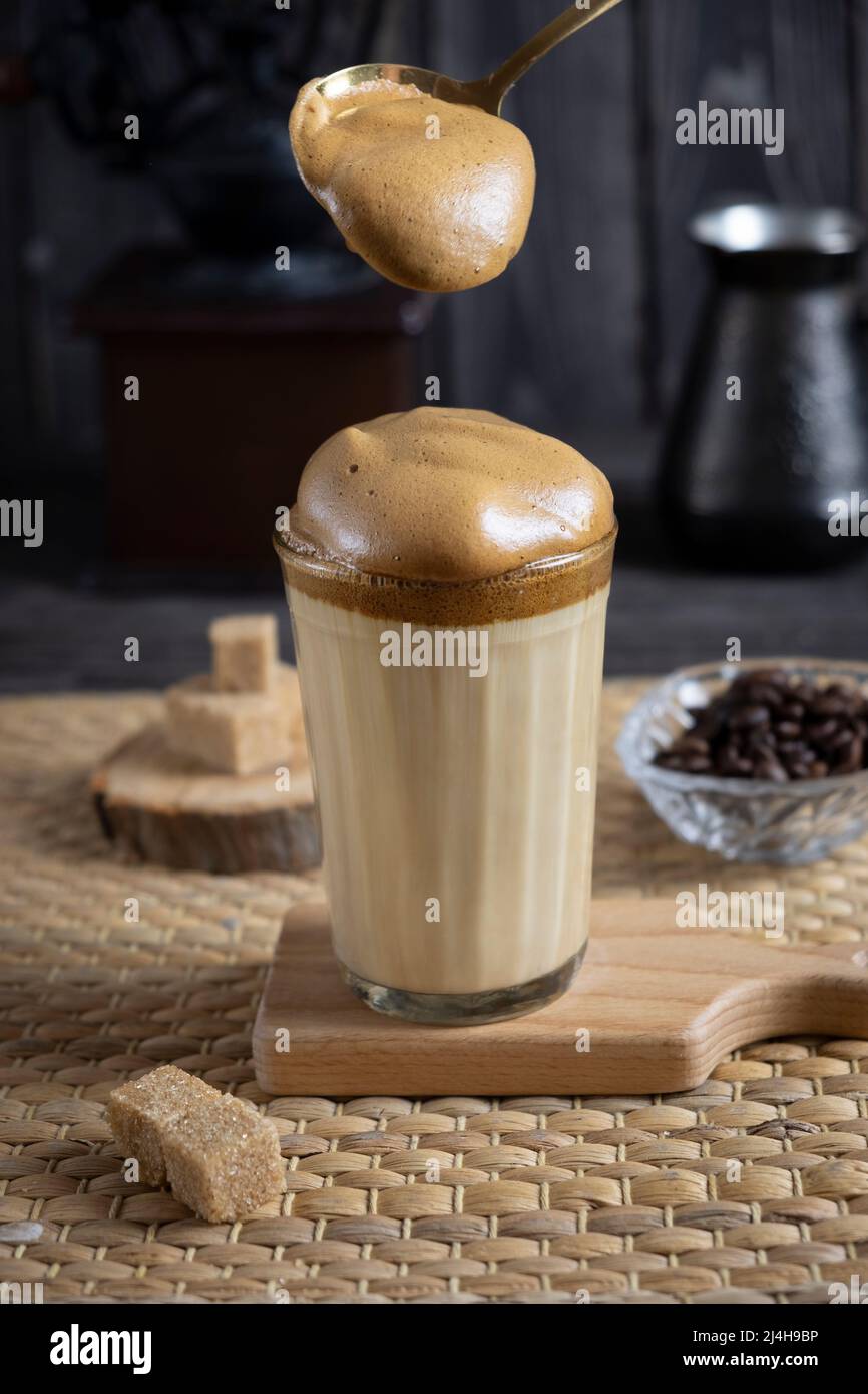 Caffè Dalgona ghiacciato, caffè cremoso alla moda montato su sfondo di legno scuro con macinacaffè, zucchero e chicchi di caffè, con cucchiaio di schiuma Foto Stock