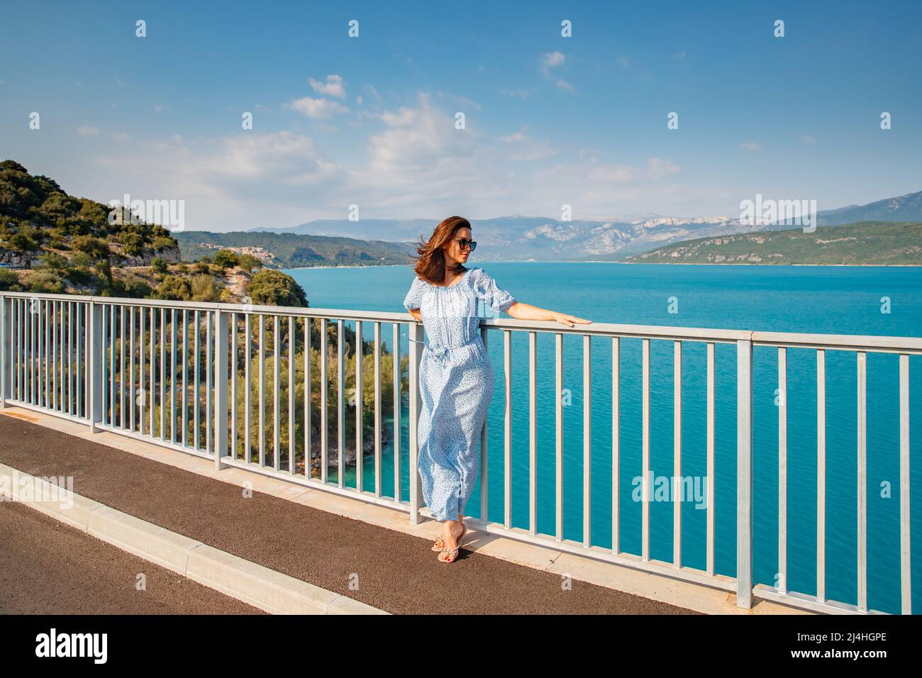La bella ragazza in un abito blu e occhiali da sole pone sul ponte, i lunghi capelli castagno, felice e sorrisi, l'acqua azzurra del lago e pendii di Foto Stock