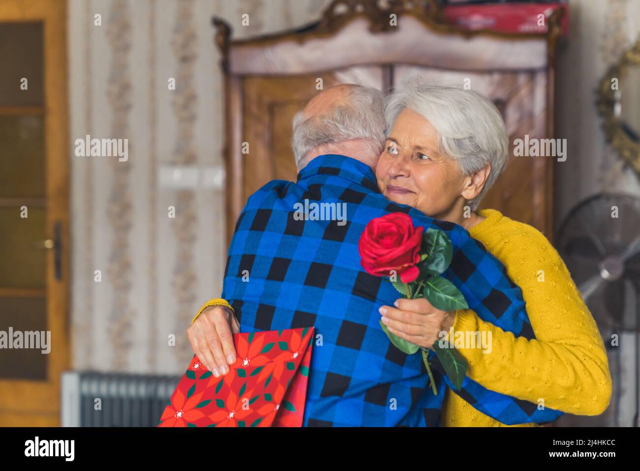 Concetto di celebrazione romantica. Anziana coppia caucasica festeggia il loro anniversario. Uomo in pensione che abbraccia la moglie dai capelli grigi che tiene una rosa rossa e una borsa regalo. Foto di alta qualità Foto Stock