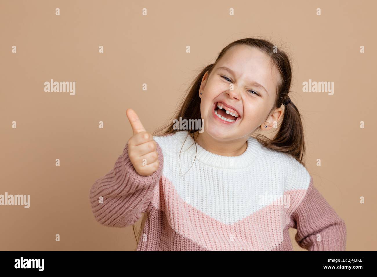 Ritratto di giovane adorabile ragazza ridente gioiosa con lunghi capelli scuri in bianco, maglione rosa in piedi, mostrando pollici segno, gesto di approvazione su Foto Stock