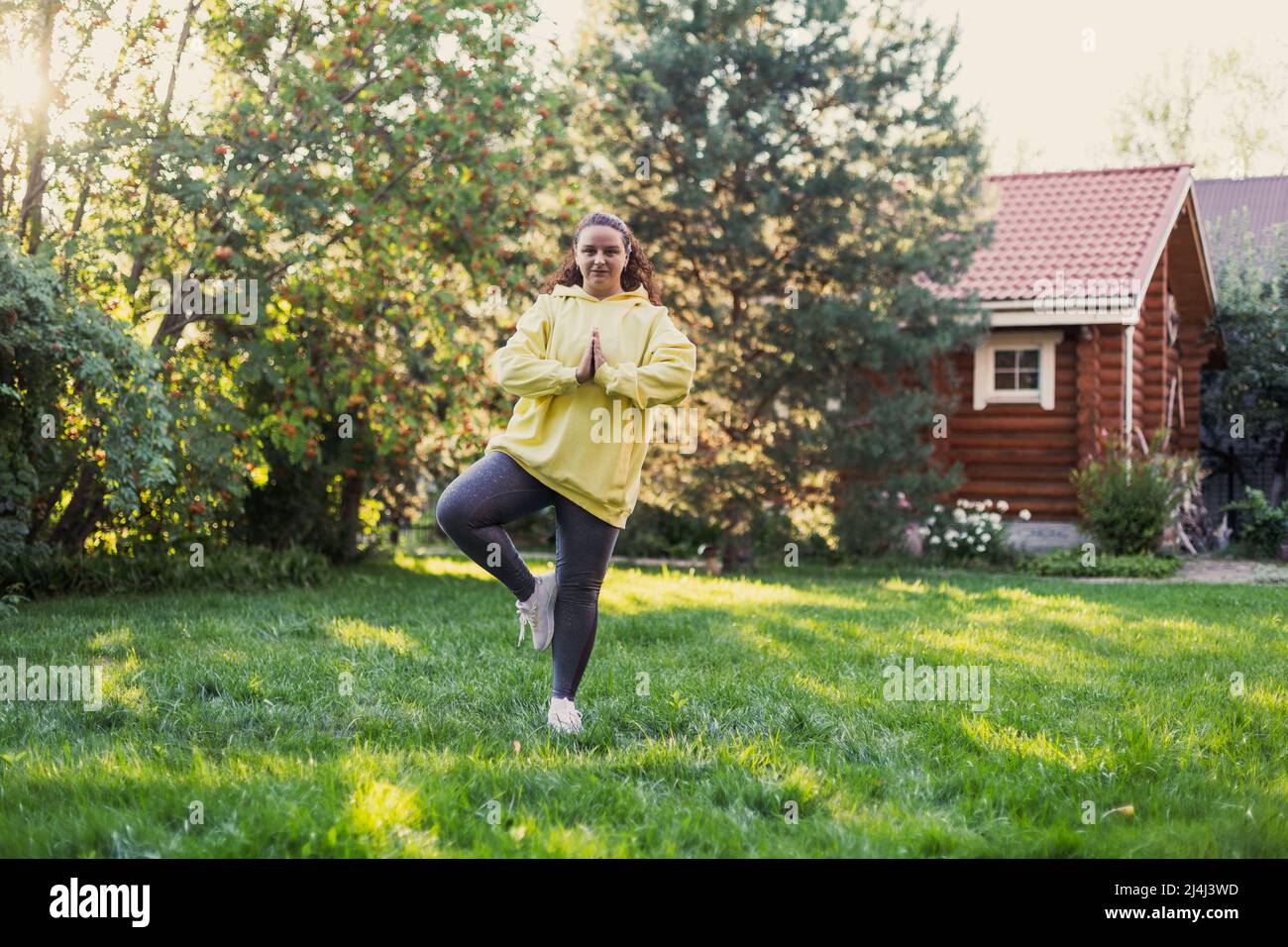 Donna che si bilancia su un piede di erba fresca facendo esercizio yoga indossare abiti sportivi guardando la macchina fotografica sul cortile con casa di campagna e alto Foto Stock